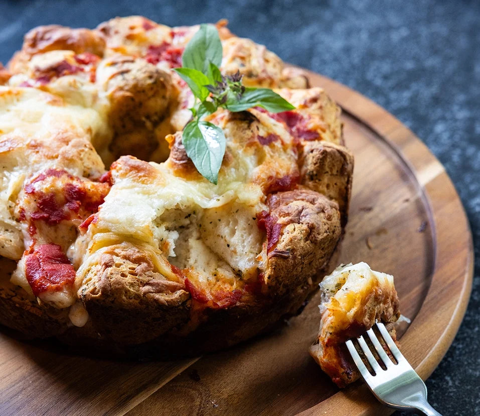 A close-up of a baked pizza pull-apart bread on a wooden board, with a fork lifting a piece and a fresh basil sprig on top.