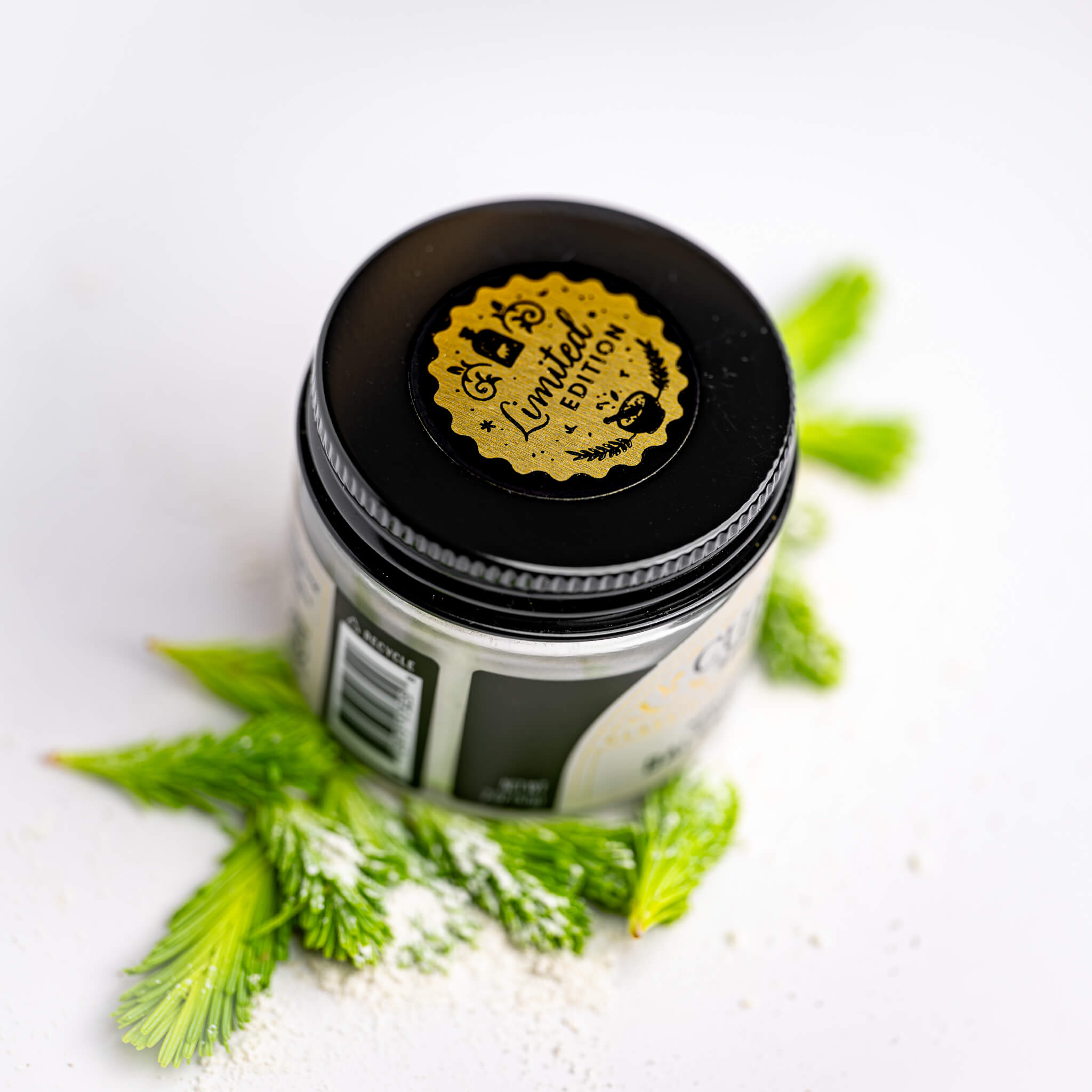 Jar of Balsam Sugar with a black lid featuring a gold limited edition label, surrounded by green fir tips on a white background.