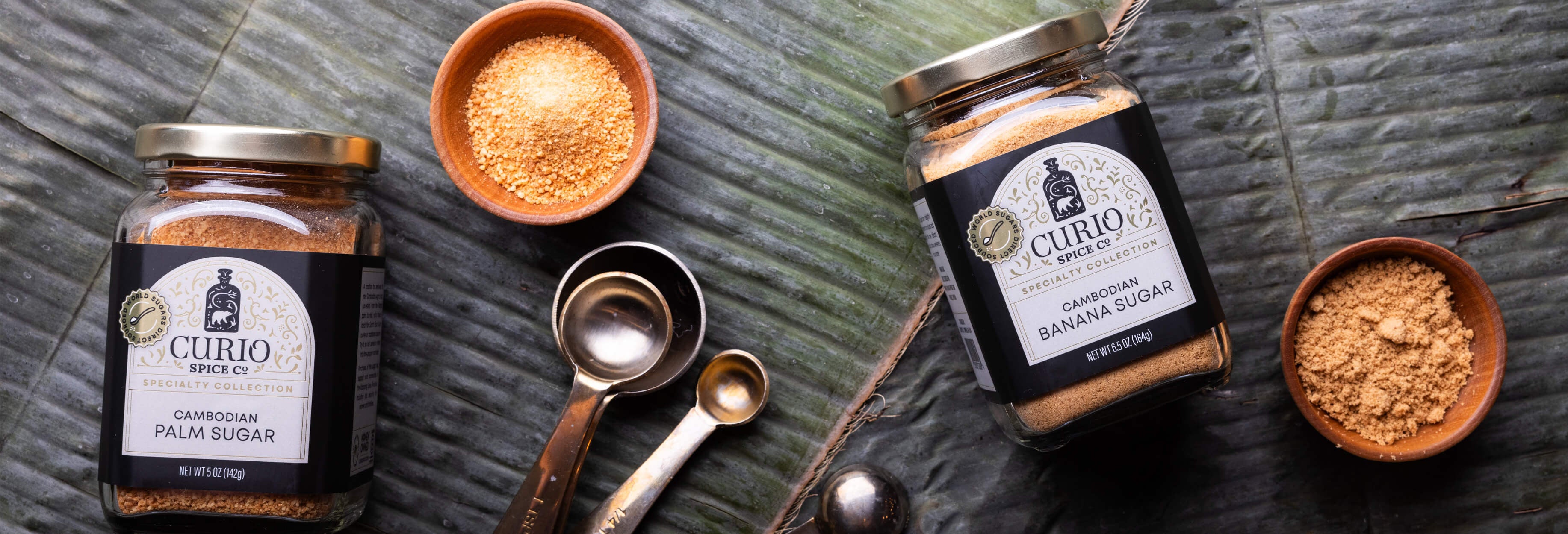 Two jars of Curio sugars with bowls and measuring spoons on banana leaves