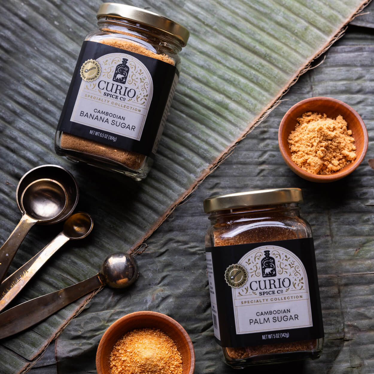 Two jars of Curio Spice Co. Cambodian banana and palm sugar on a textured surface with measuring spoons.