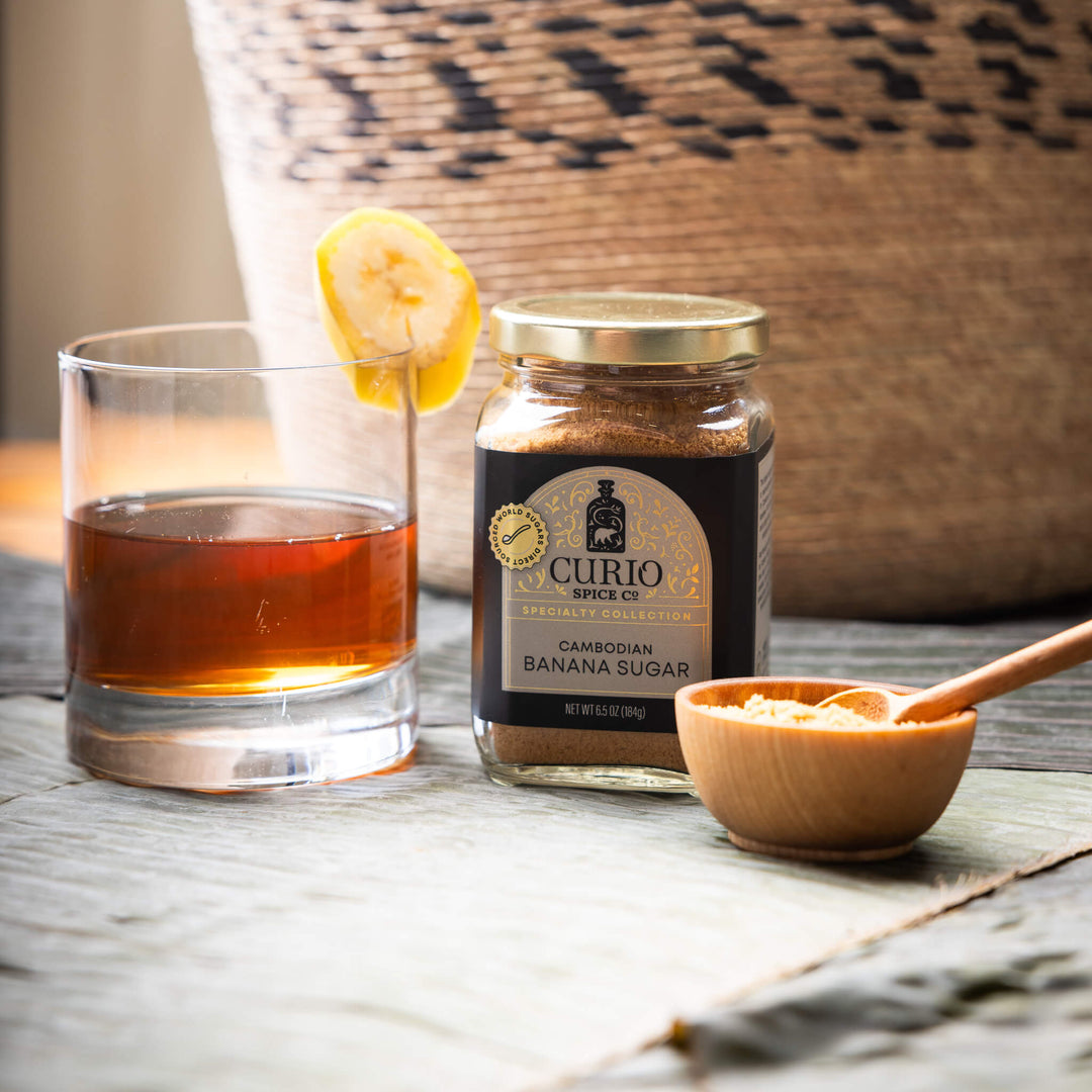 Glass of amber liquid with a lemon slice, jar of Curio Spice Co. Cambodian Banana Sugar, and wooden spoon on a wooden surface.