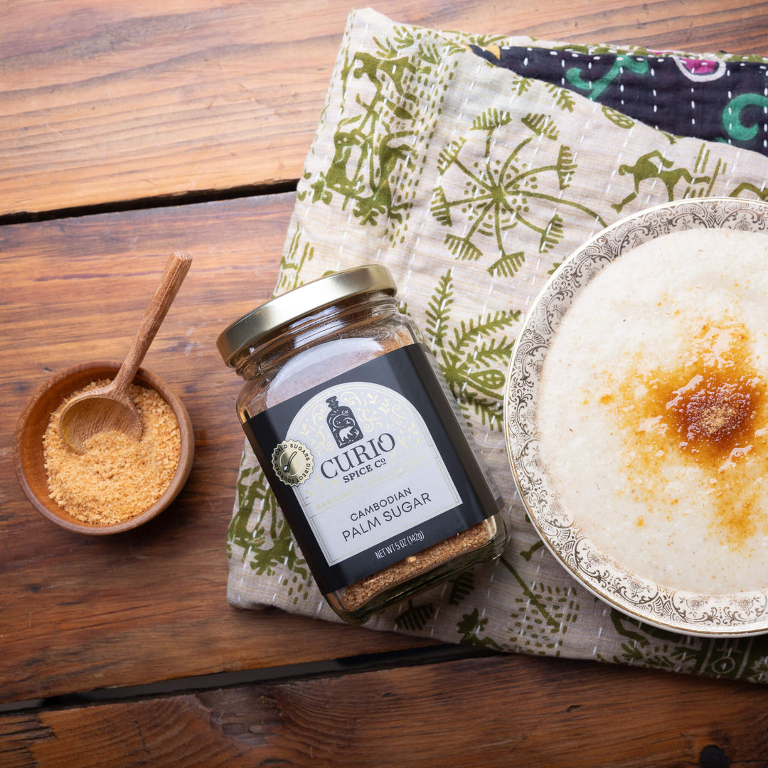 Jar of Curio Specks Cambodian Palm Sugar on a wooden table with a bowl of sugar and a plate of food.