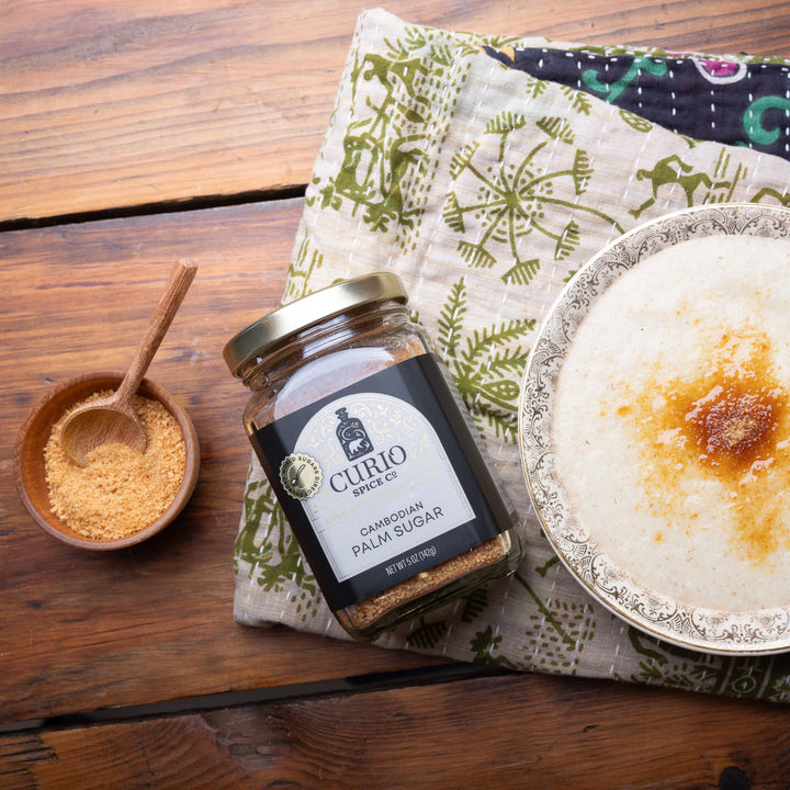 Jar of Curio Specks Cambodian Palm Sugar on a wooden table with a bowl of sugar and a plate of food.