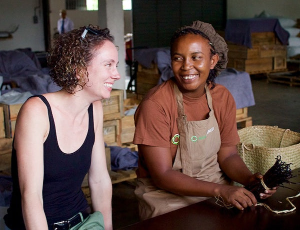Two women sitting together, one wearing a black tank top and the other in a brown apron, in a room with wooden crates and baskets.