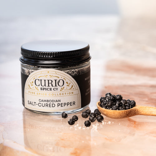 Jar of Cambodian Salt-Cured Pepper from Curio Spice Co. with a wooden spoon of pepper on a light background