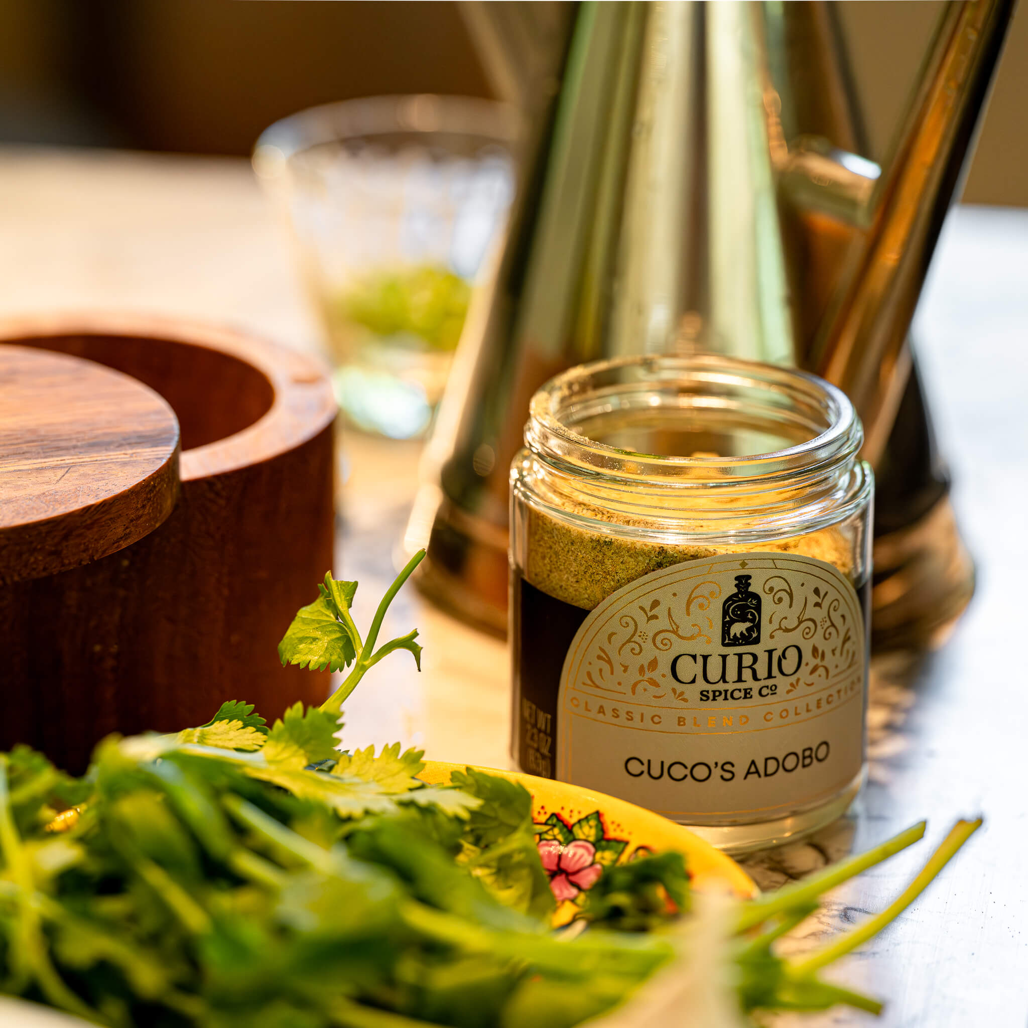 Jar of Curio Spice Co. adobo on a table with herbs and a blurred background