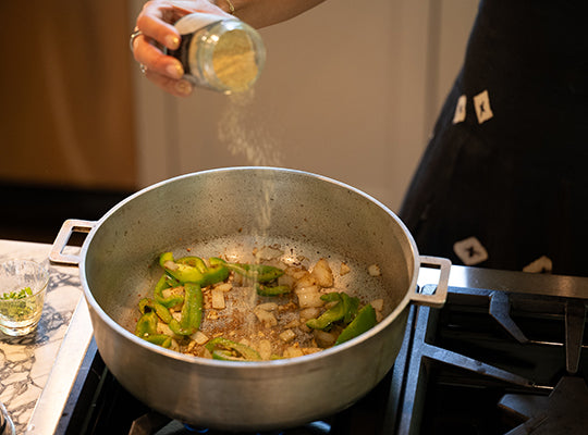 Person seasoning vegetables in a pan for Cuban sofrito on a stove.