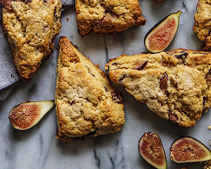 Several triangular fig scones are arranged on a light marble surface with sliced figs placed around them.