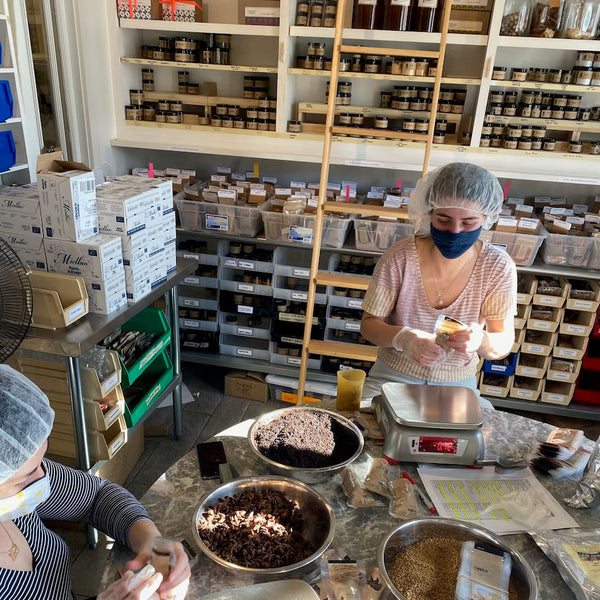 Two masked people working in a workshop with shelves of jars and boxes.