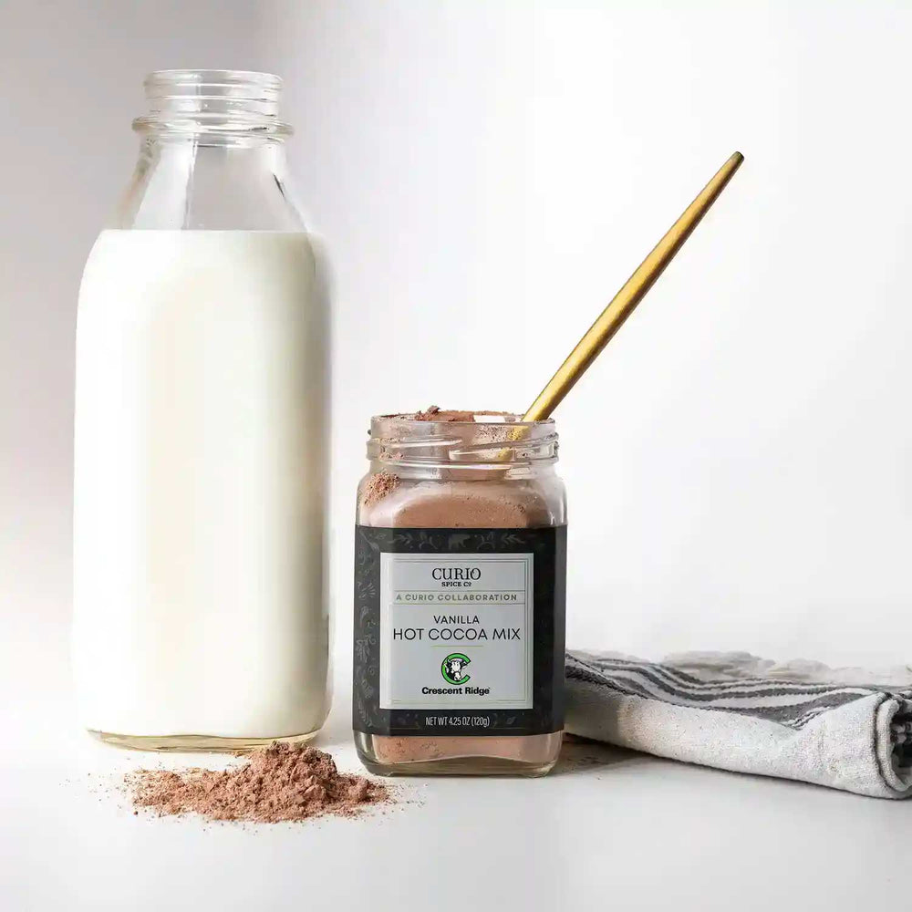 Open glass jar of Vanilla Hot Cocoa Mix next to a scatter of the mix, a jug of milk, and a towel.