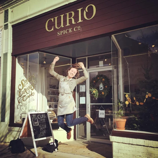 Claire Cheney jumping in front of a store named 'Curio Spice Co' with a chalkboard sign that says "Now Open!"