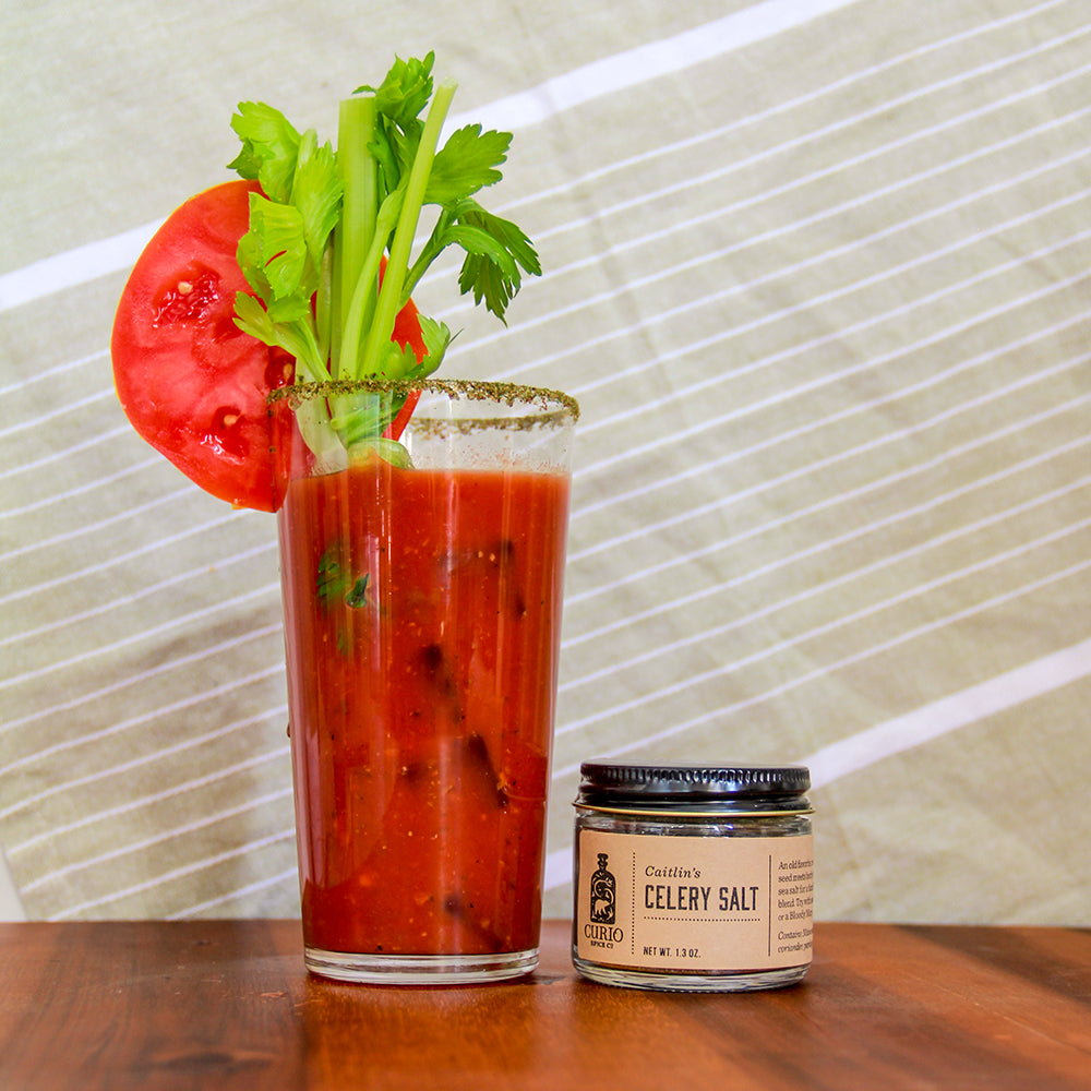 Bloody mary with celery and tomato garnish next to a jar of celery salt on a wooden surface.
