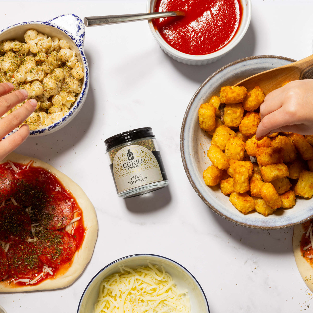 An overhead flat-lay of pizza preparation on a white surface, featuring a jar of Curio Spice Pizza Tonight! seasoning, bowls of cheese, tomato sauce, and golden tater tots, with hands reaching in.