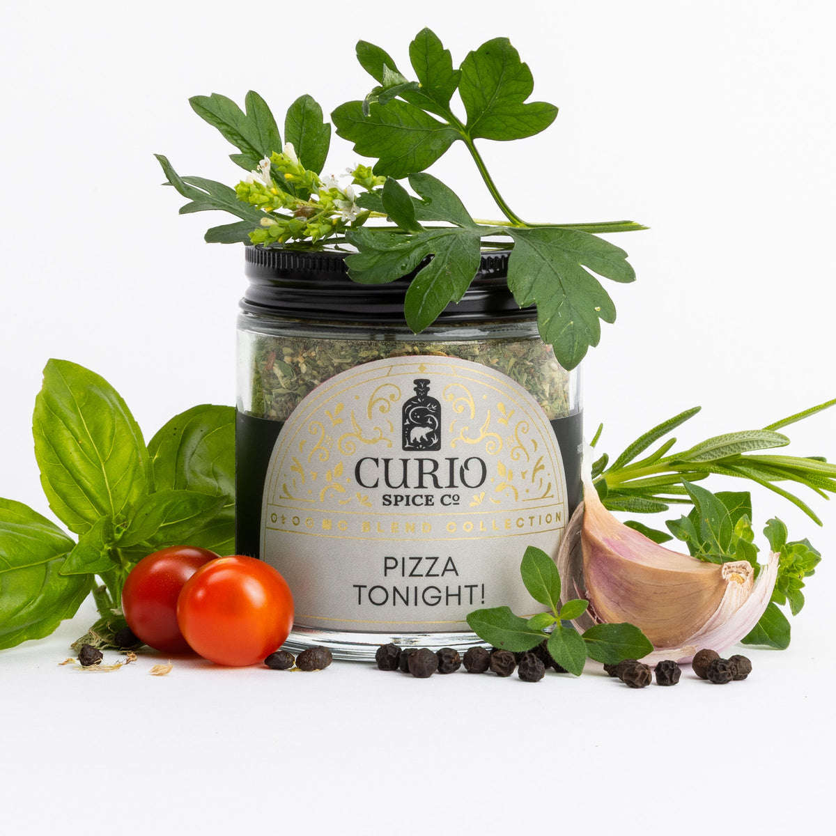A jar of Curio Spice Co. 'Pizza Tonight!' organic spice blend, styled with fresh green herbs, two red cherry tomatoes, a clove of garlic, and black peppercorns on a clean white background.