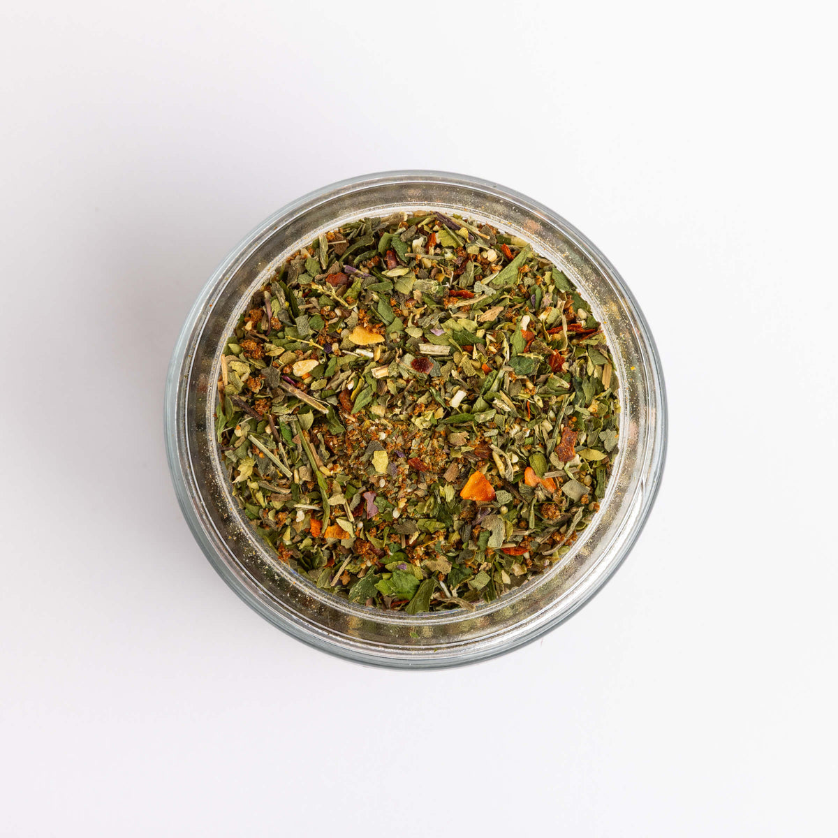 Top-down view of a clear glass jar filled with a loose spice blend containing green, brown, and orange dried botanicals, set against a plain white background.