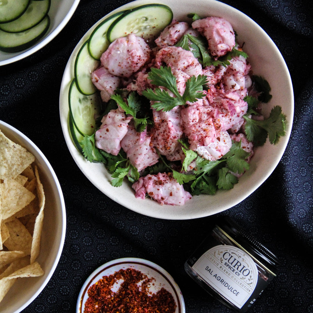 Bowl of pink ceviche with cilantro, surrounded by sliced cucumbers, tortilla chips, and a jar of Curio Spice Co Sal Agridulce on a dark surface.