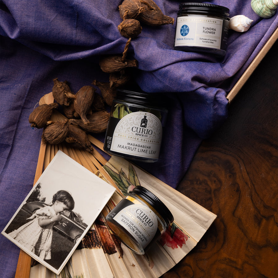 Jars of Curio spices on a wooden board with dried flowers and a vintage photo on a purple fabric background.