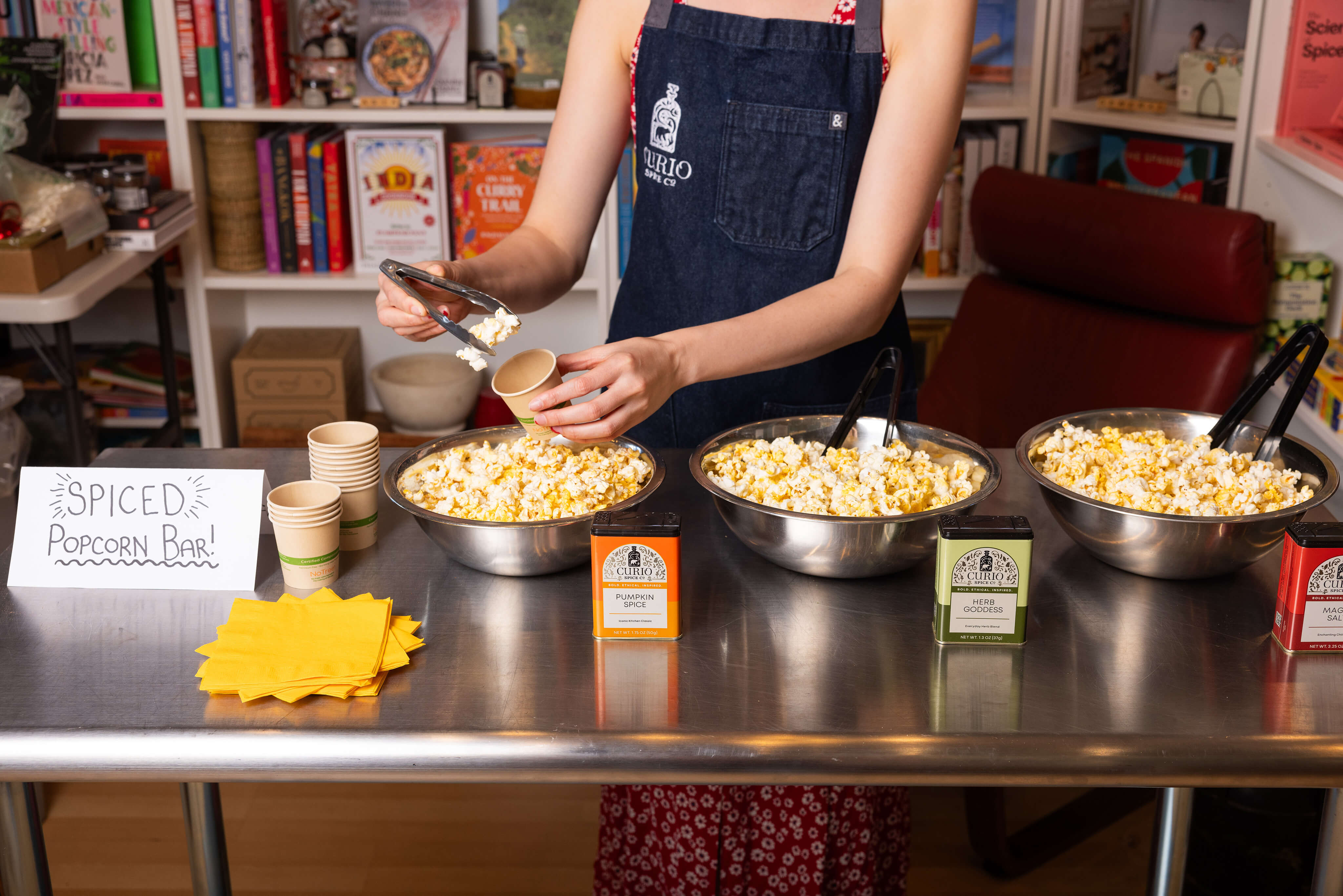 A spiced popcorn bar with hands putting some popcorn into a cup with tongs. The popcorn uses Curio's Everyday Blends.