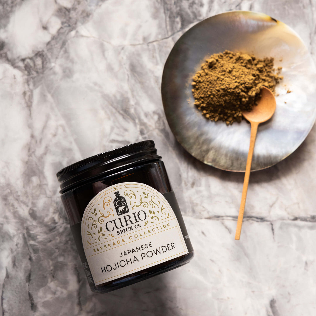 Jar of Curio Spice Co. Japanese Hojicha Powder on a marble surface with a spoonful of powder.