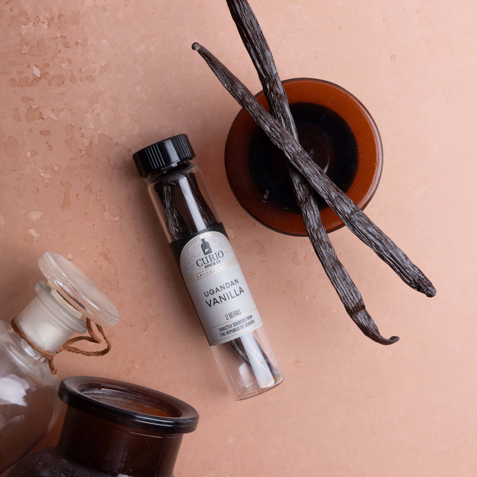 A small clear glass vial lies on a peach surface, surrounded by vanilla beans and amber glass bottles.