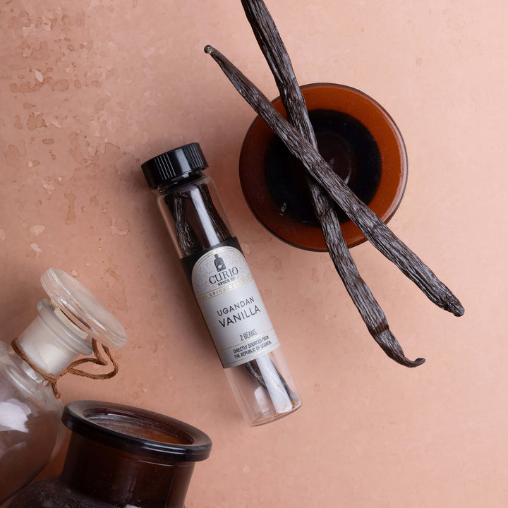 A small clear glass vial lies on a peach surface, surrounded by vanilla beans and amber glass bottles.