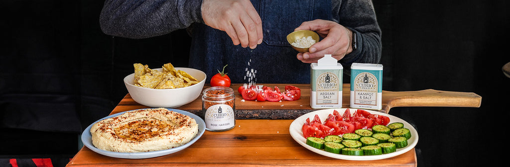 Slideshow image of Rose Harissa, Aegean Salt, and Kampot & Salt being sprinkled on hummus and vegetables.