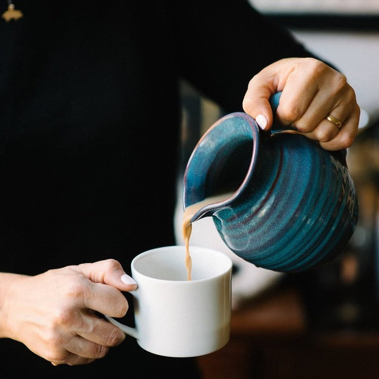 Image of a person pouring Uncommon Chai from a blue ceramic jug into a tea cup.